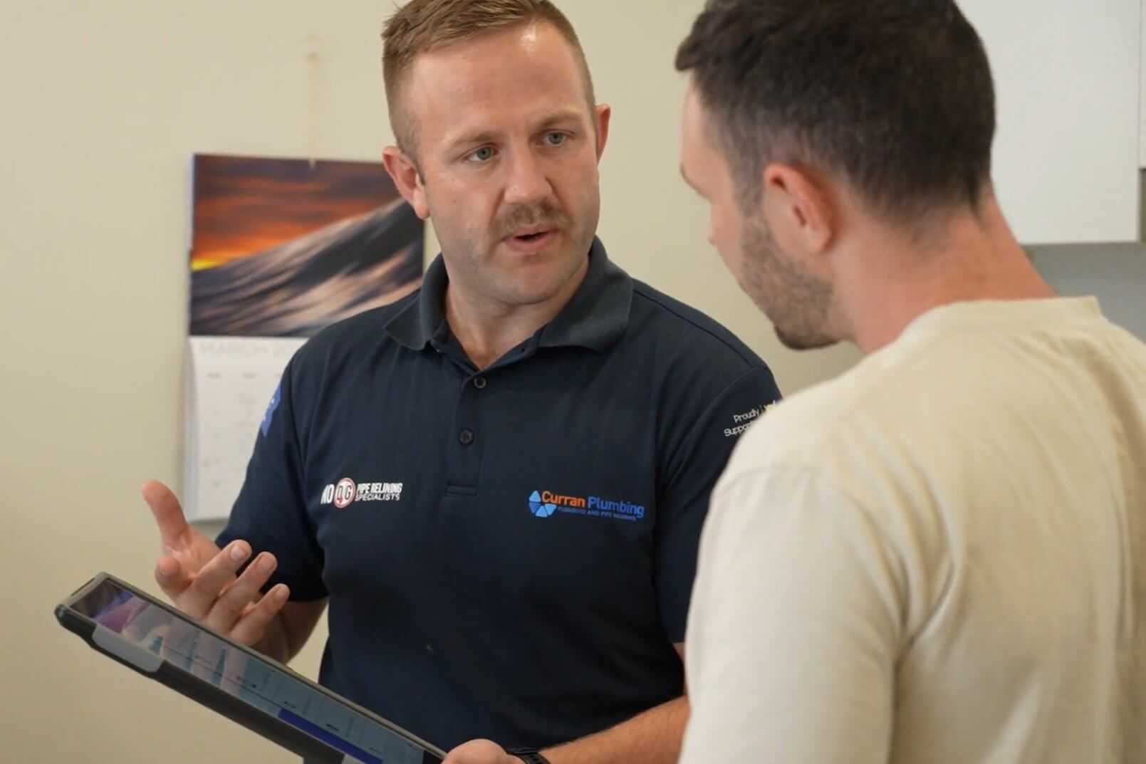 Plumber wearing navy top showing quote on tablet to client Plumber wearing navy top showing quote on tablet to client